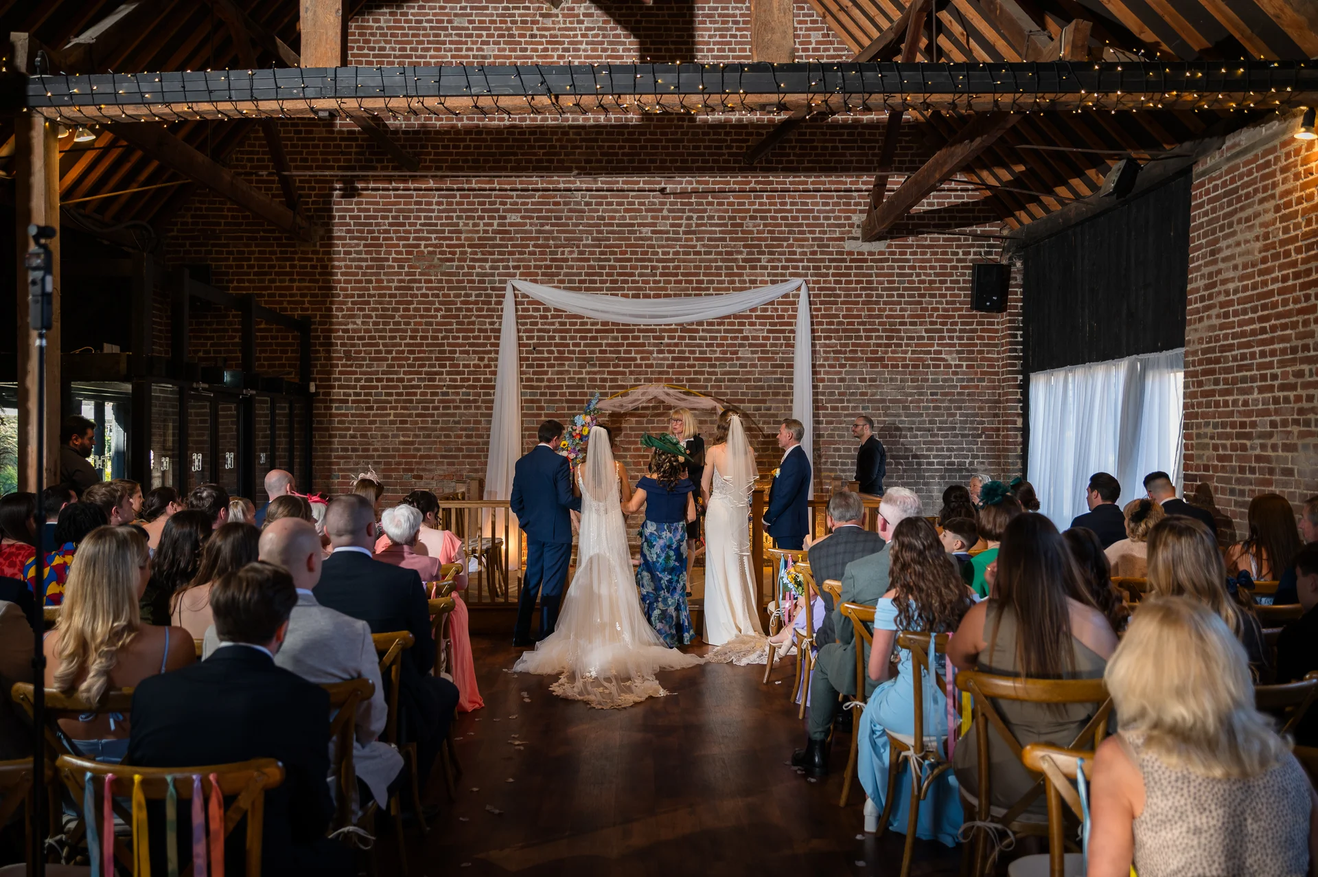 A wide shot of a wedding ceremony inside a rustic brick barn with wooden rafters, showing the couple and guests seated on wooden chairs.