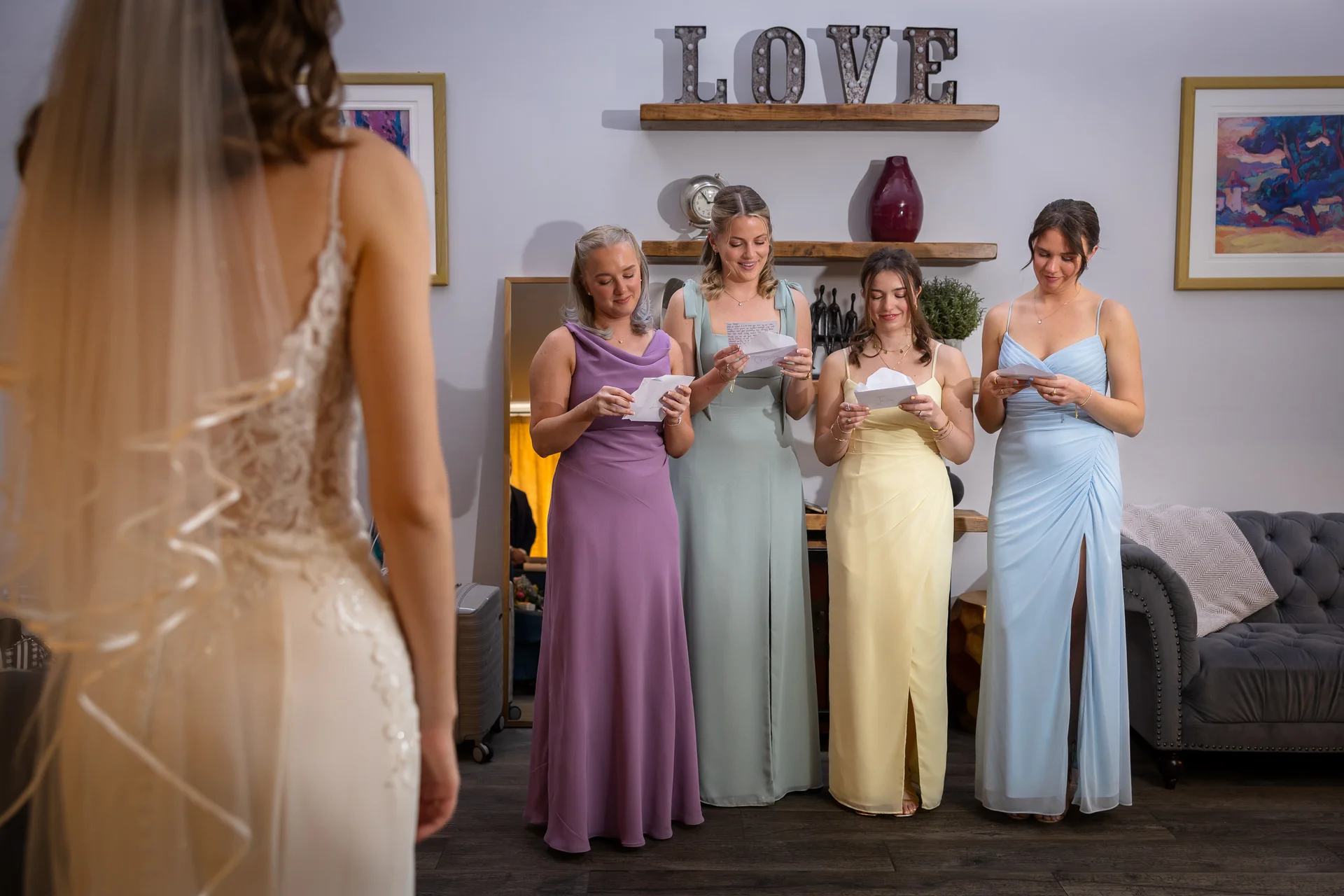 A bride in a lace wedding dress with a veil stands in the foreground, turning to watch her four bridesmaids read heartfelt letters aloud.