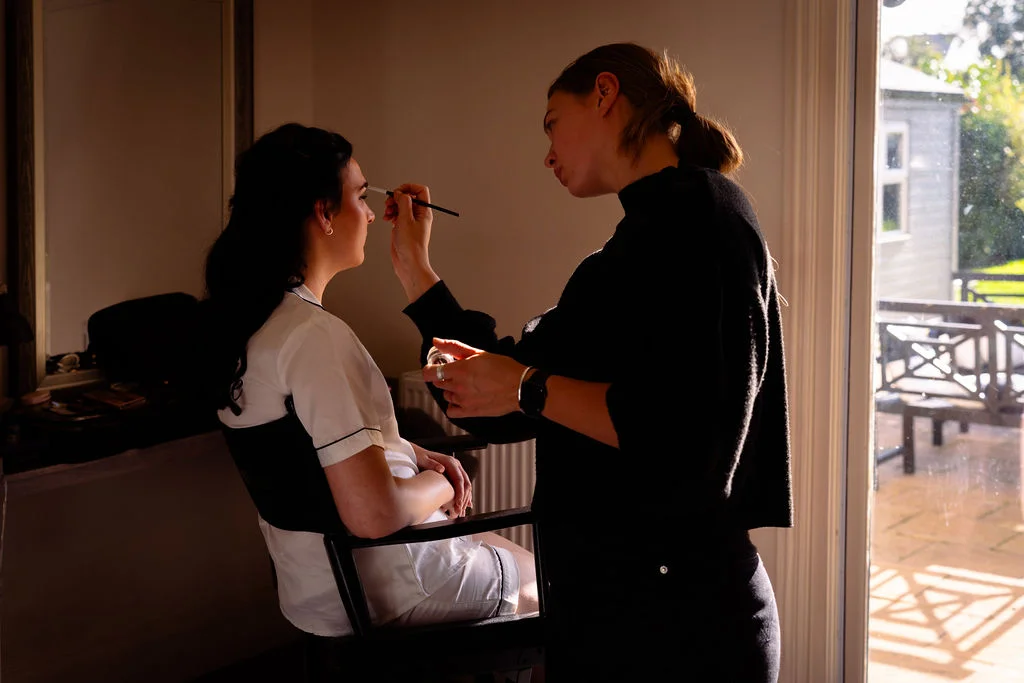 A professional makeup artist carefully applies eyeliner to a bride wearing a white getting-ready robe, bathed in warm backlit window light.