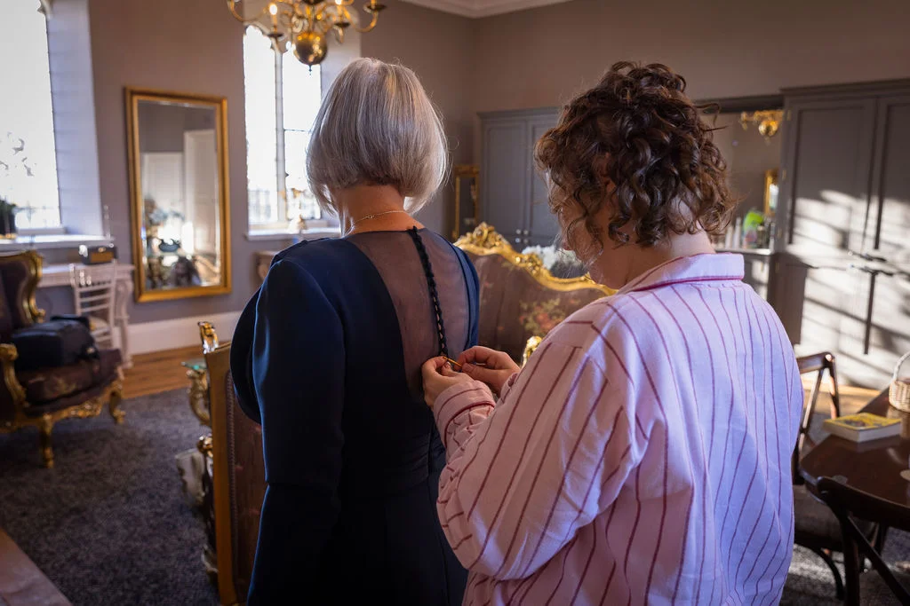 A woman in striped pyjamas carefully fastens the buttons on the back of an elegant dark blue dress during morning wedding preparations.