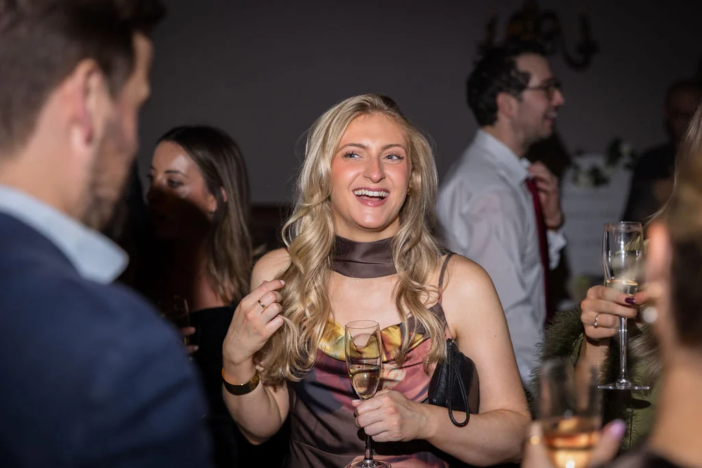 A smiling blonde guest holding a glass of champagne shares a joyful moment while mingling with other attendees during the evening wedding reception.