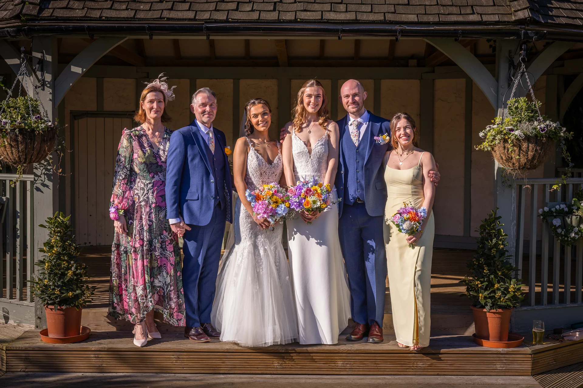 A happy bridal party and family group pose together outdoors on a wooden deck in front of a rustic gazebo on a sunny wedding day.