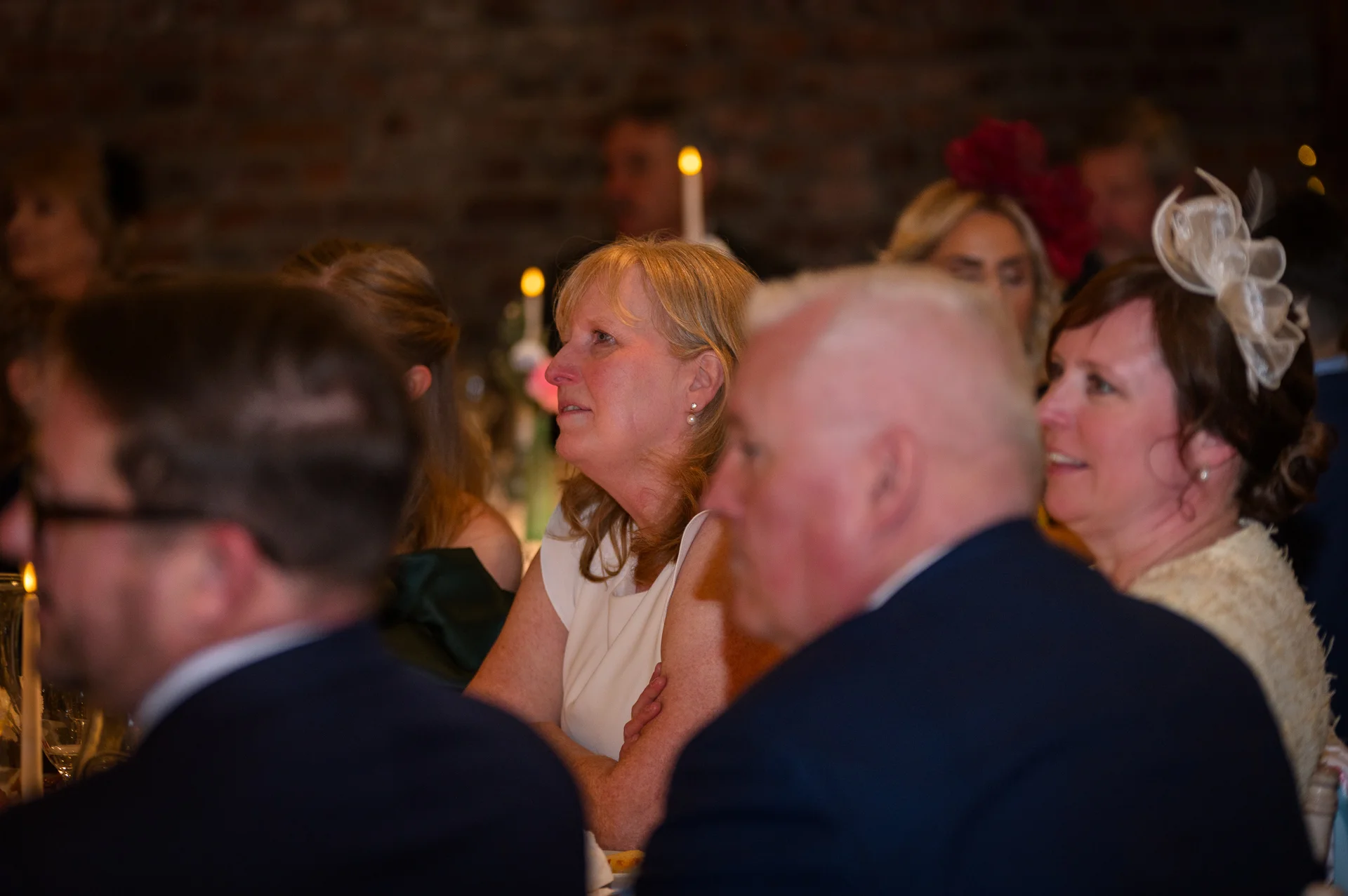 Wedding guests seated at tables inside a rustic venue with exposed brick walls, watching speeches and reacting with smiles and laughter.