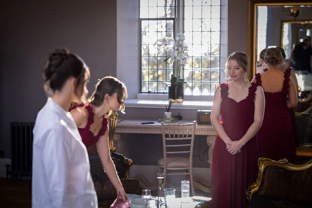 A bridesmaid in a burgundy sweetheart gown stands in front of a mirror while getting ready in a sunlit room.