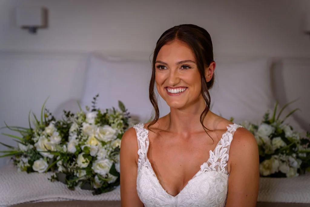A smiling bride wearing a lace v-neck wedding dress sits indoors with white floral bouquets arranged behind her.