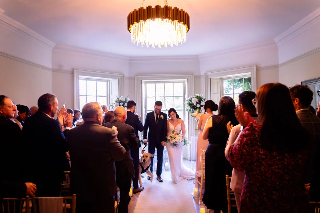 Newlyweds walk down the aisle together accompanied by their dog, with cheering guests applauding on either side in a bright, elegant room illuminated by a large chandelier.