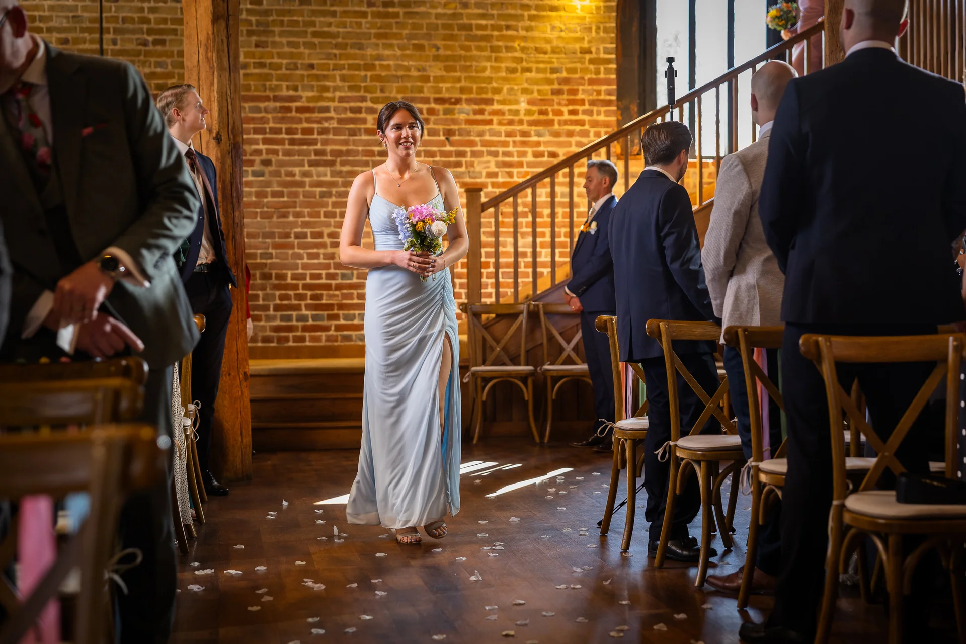 A smiling bride in a light blue slip dress with a thigh split walks down the aisle strewn with petals inside a rustic stone and brick barn.