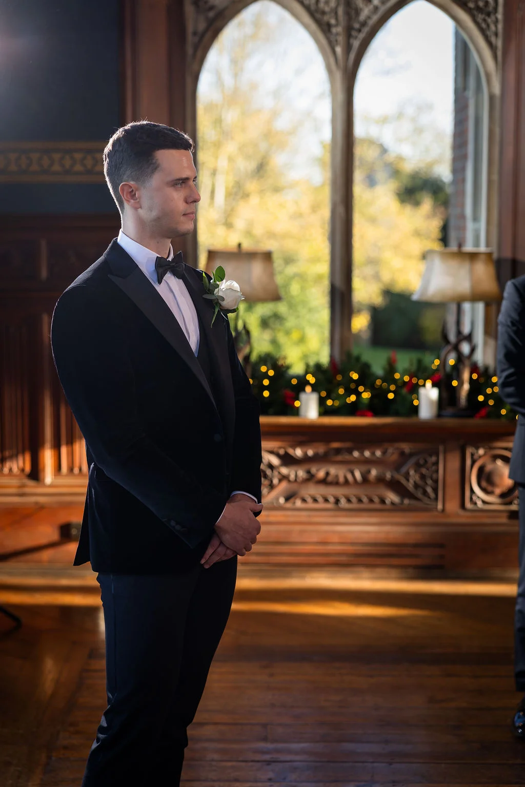 A handsome groom in a black velvet tuxedo waits attentively at the altar inside a wood-panelled venue illuminated by soft window light.