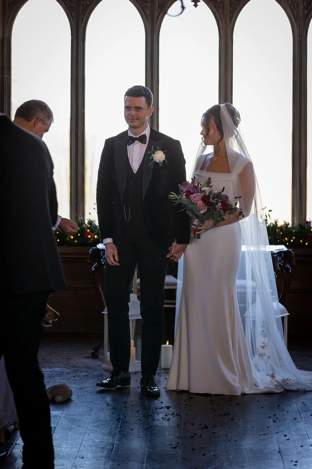 A bride in a sleek crepe gown and embroidered veil stands hand in hand with a groom in a black velvet tuxedo before tall arched windows during their wedding ceremony.