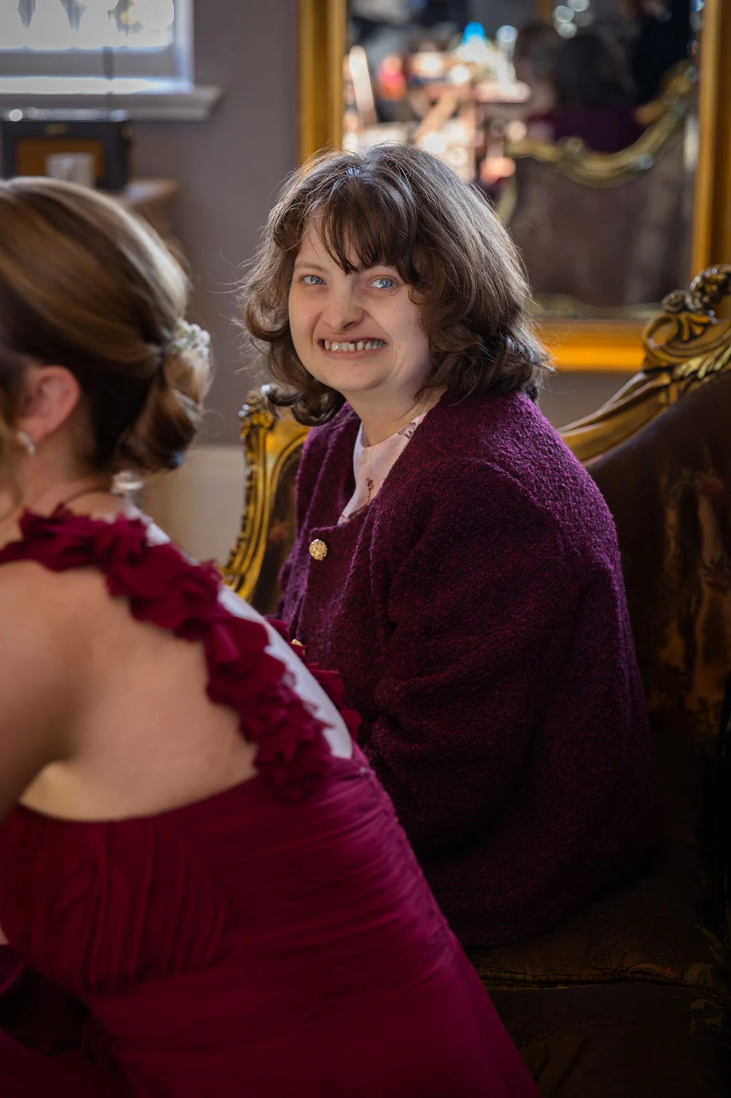 A smiling guest wearing a textured purple jacket sits on a decorative armchair while looking over her shoulder towards another person during wedding preparations.