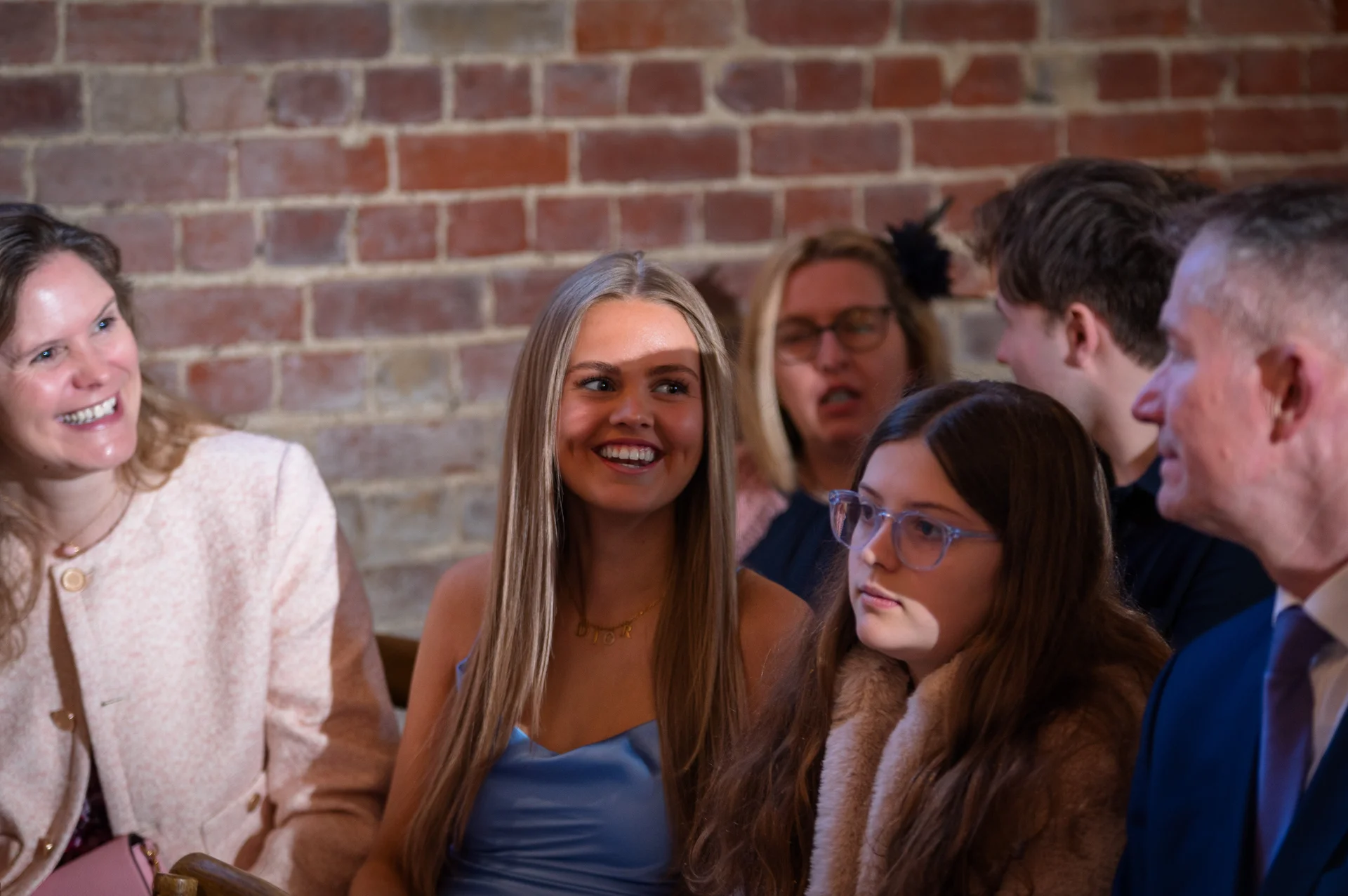 A candid group of wedding guests smiling and watching events unfold in a room with an exposed brick wall.