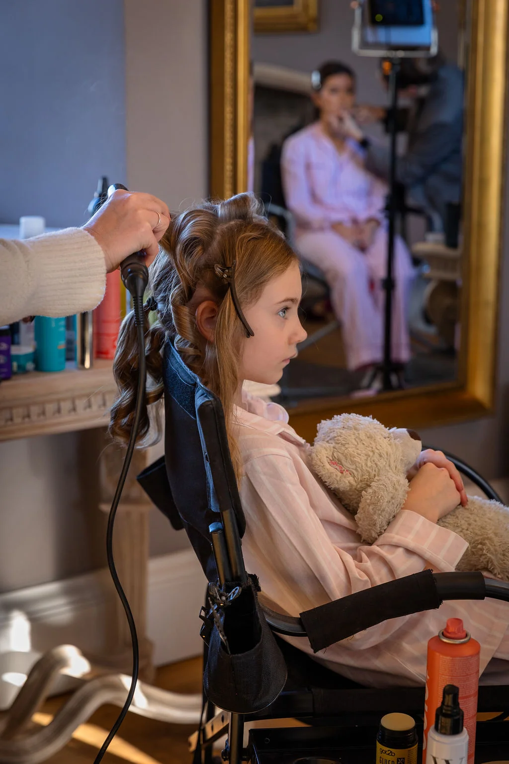 A young flower girl in striped pyjamas sits in a styling chair holding a teddy bear while getting her hair curled before the wedding.