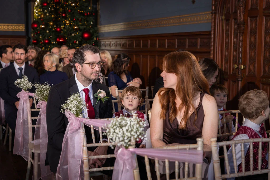 Wedding guests seated in a wood-panelled room decorated with Christmas trees and chairs adorned with pink sashes and gypsophila.