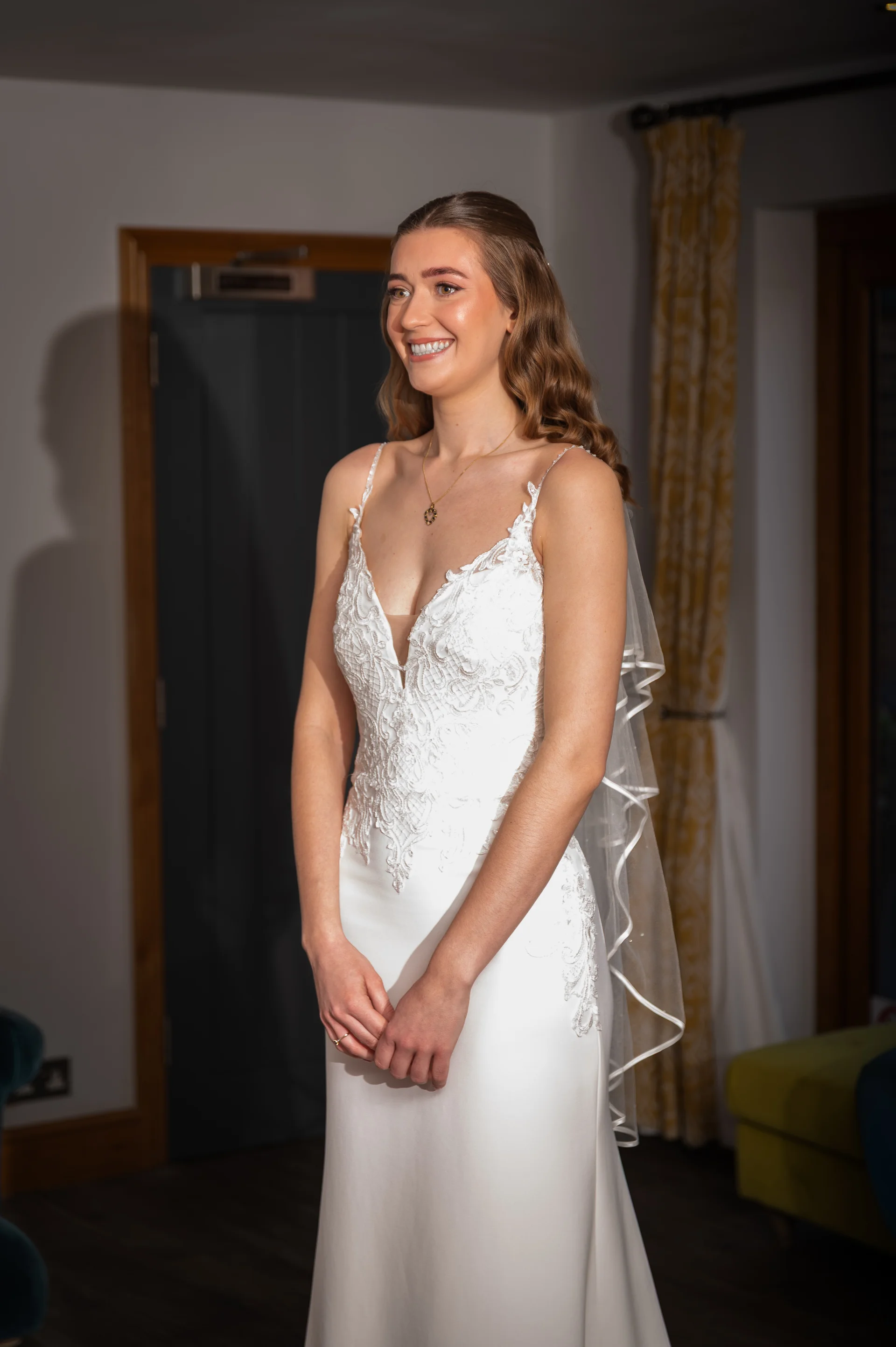 A smiling bride wearing a crepe wedding gown with a lace bodice, spaghetti straps, and a ribbon-edged veil poses indoors during bridal preparations.