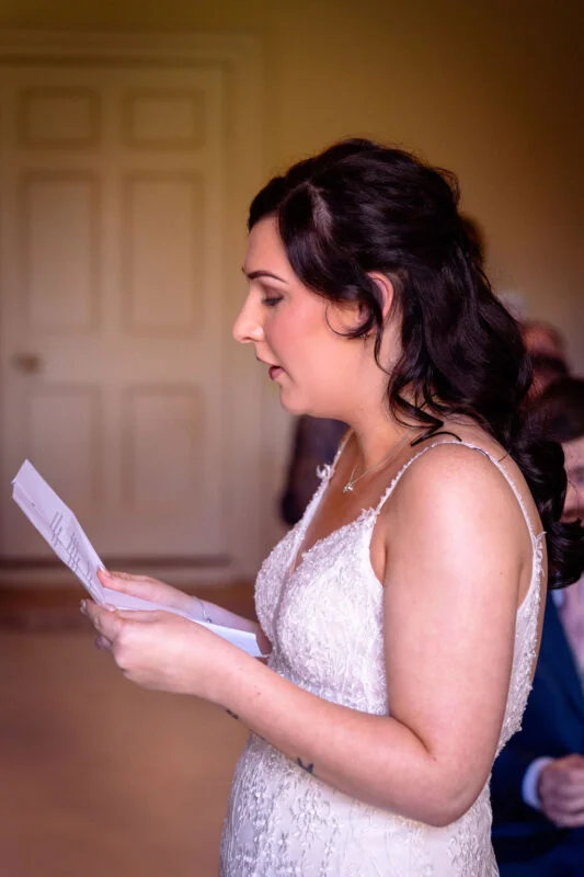 A profile shot of a bride reading her personal vows from a paper during an indoor wedding ceremony, wearing a lace V-neck dress with spaghetti straps.