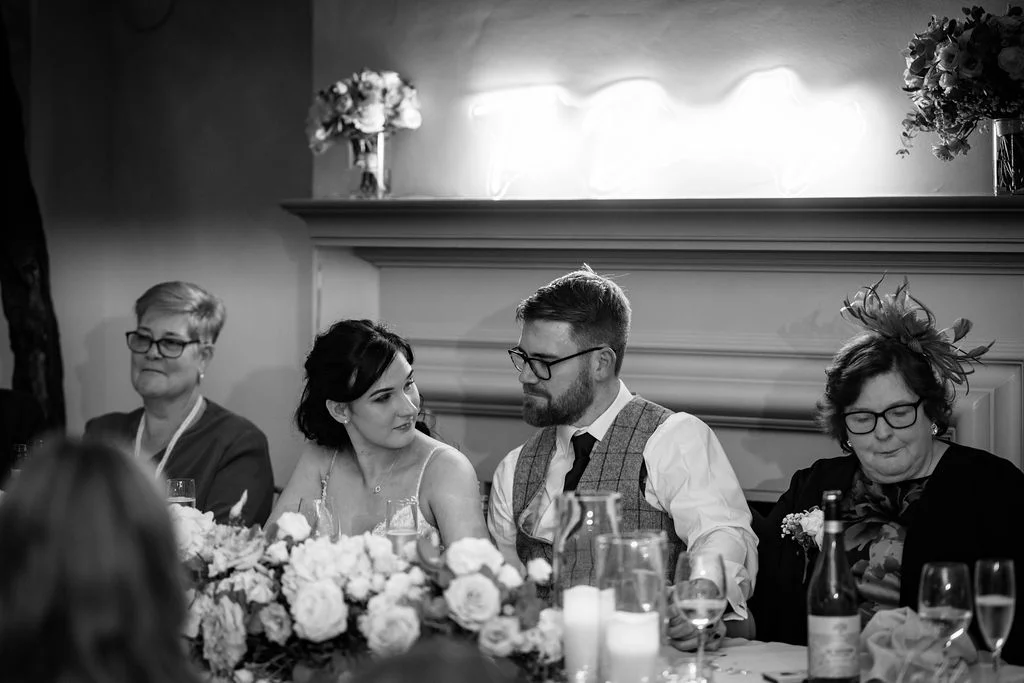A newlywed couple shares a tender glance at the top table during wedding speeches, framed by a neon sign and floral arrangements.