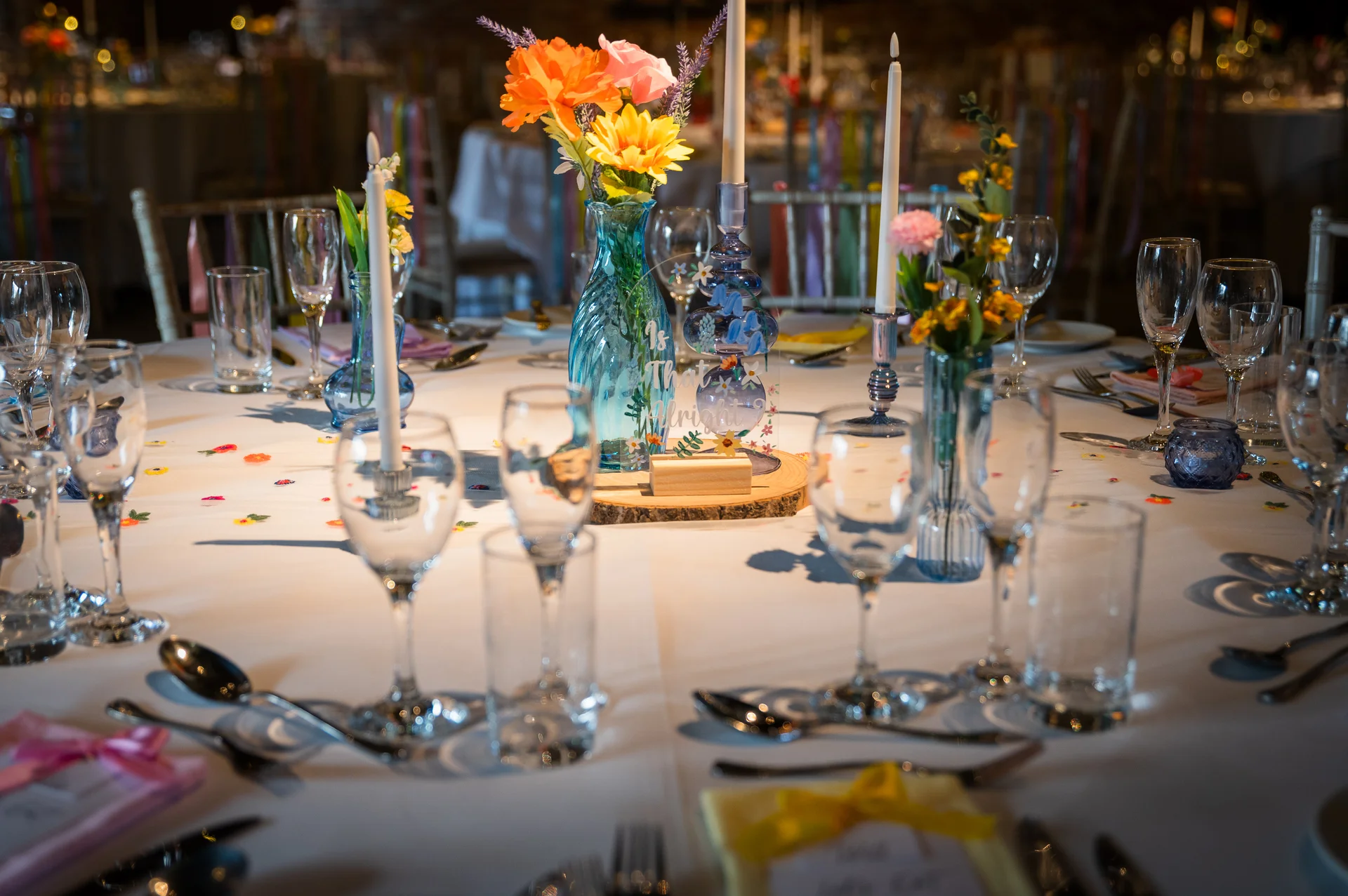 A brightly decorated wedding tablescape features colourful floral centerpieces in blue glass vases, tall taper candles, and delicate glassware on a white linen tablecloth.