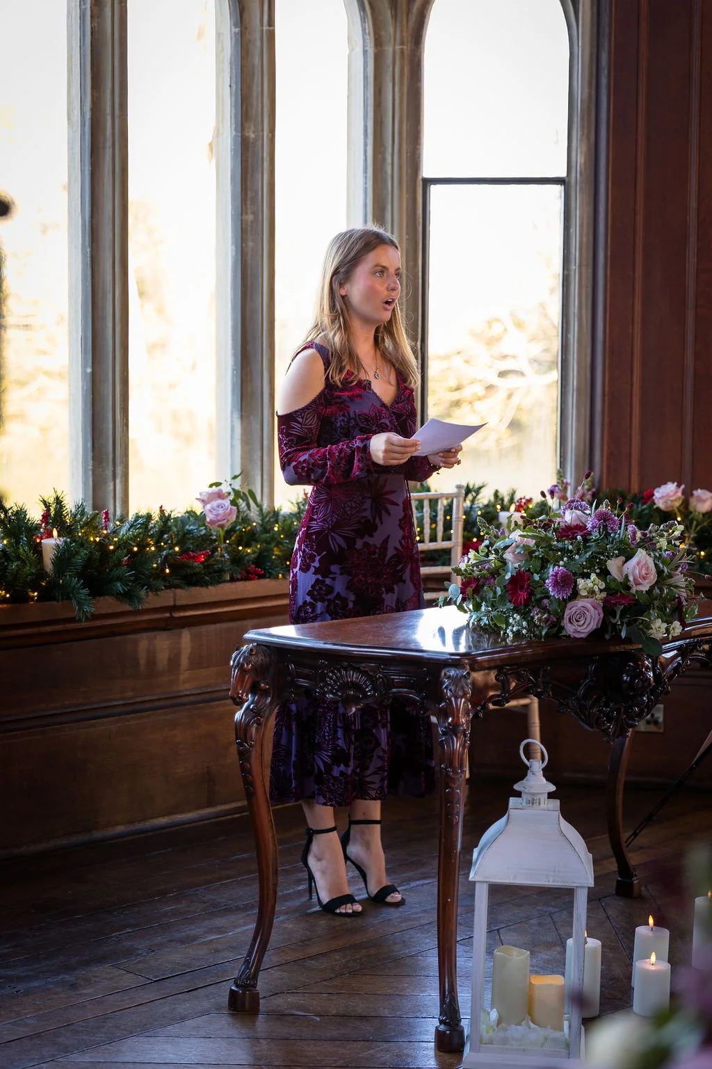 A bridesmaid delivers a speech standing beside an ornate wooden table adorned with floral arrangements and candles in front of tall windows.