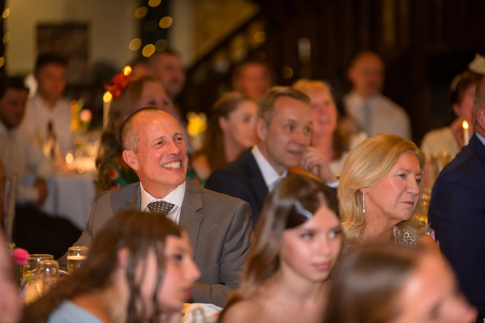 A smiling guest in a grey suit and patterned tie reacts joyfully during the wedding speeches surrounded by seated guests.