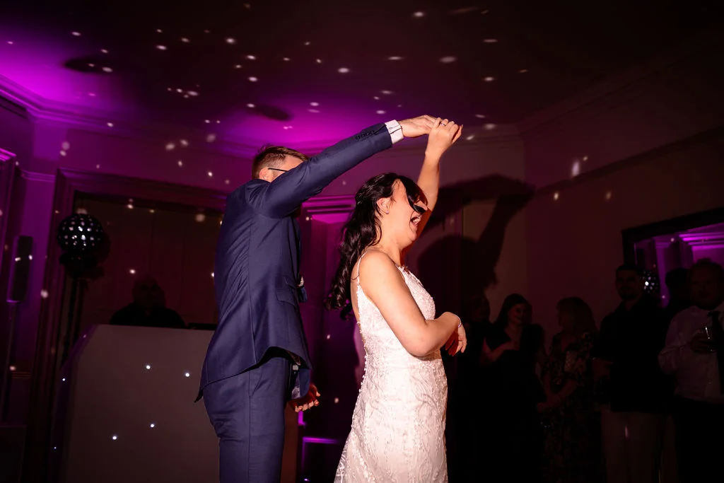 The newlyweds share a joyful moment during their first dance under dramatic purple lighting and scattered disco ball reflections.