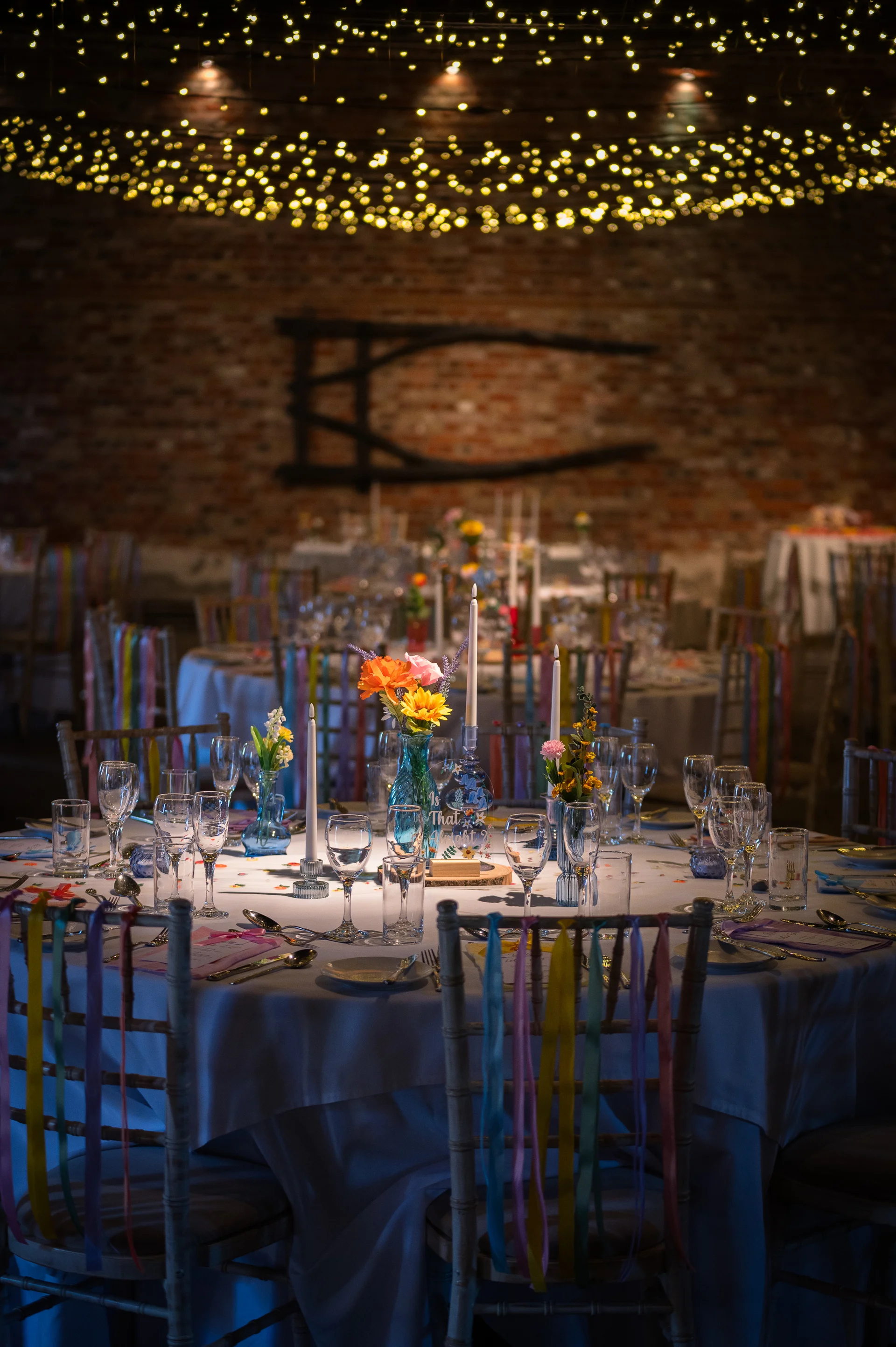 A rustic barn wedding reception featuring round tables adorned with colourful wildflowers, taper candles, and chairs decorated with hanging pastel ribbons beneath a canopy of fairy lights.
