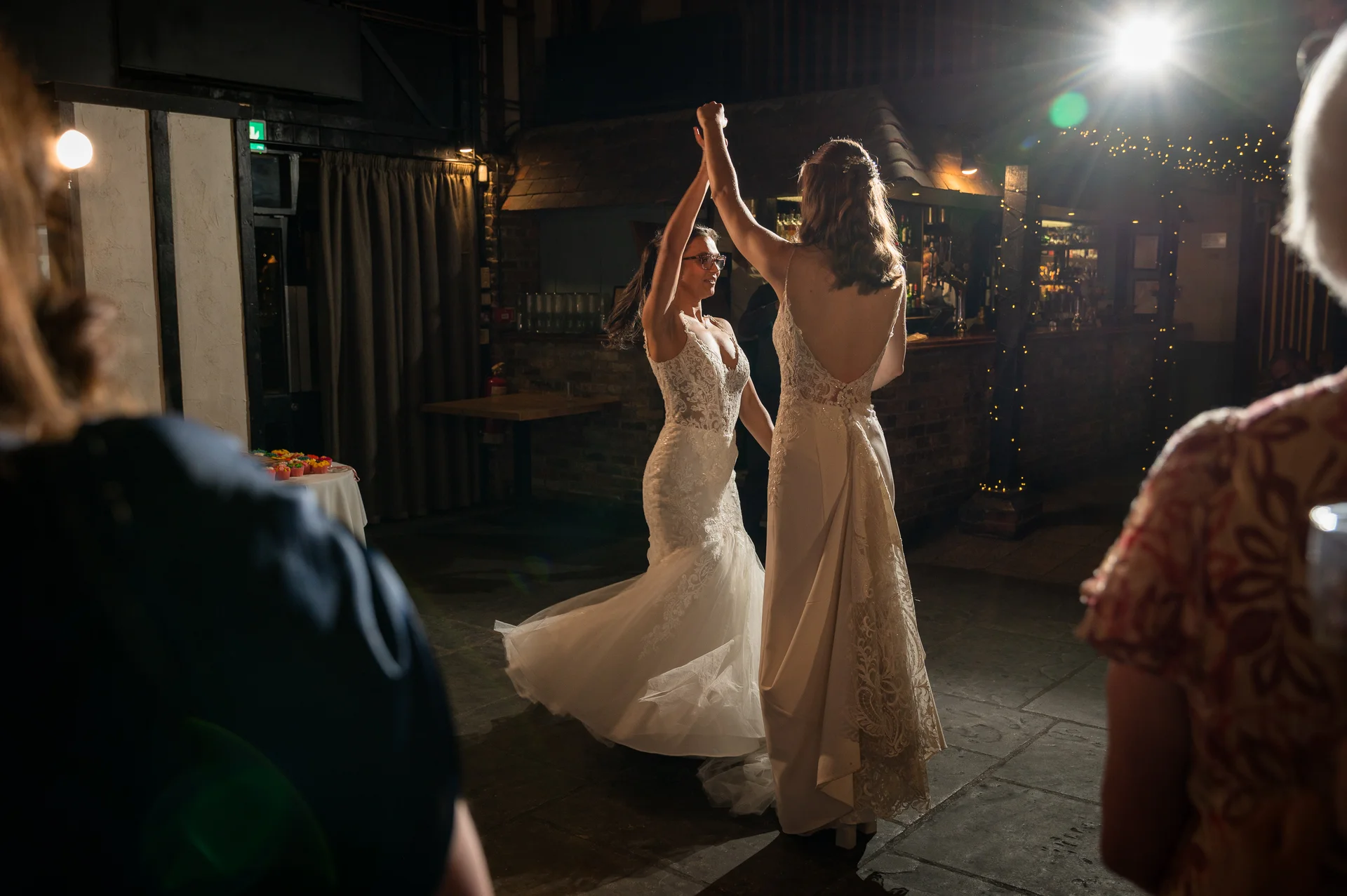 Two brides share their first dance together in a dimly lit rustic pub venue with stone flooring and string lights overhead.
