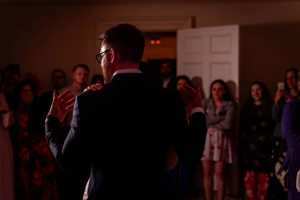 A groom in a dark suit dances with his partner during the evening reception, viewed from behind as wedding guests watch on in warm lighting.