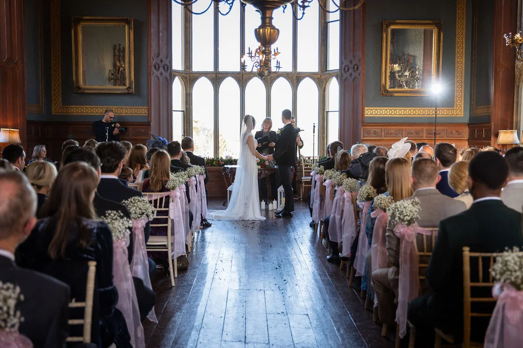 A wide shot of a wedding ceremony inside a grand manor house with guests seated along an aisle decorated with baby's breath floral arrangements.