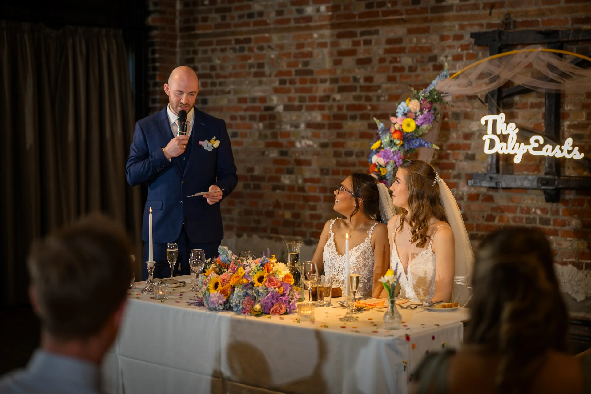 A guest delivers a speech with a microphone while two brides in lace wedding dresses listen attentively at the top table decorated with colourful floral arrangements.