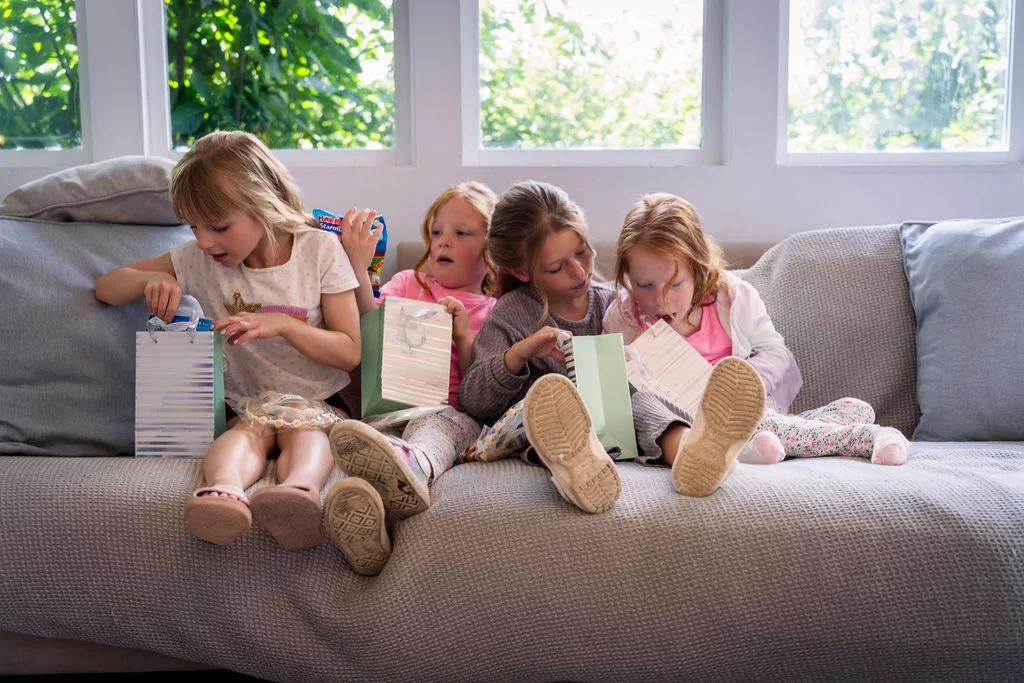 Four young flower girls sit together on a grey sofa opening gift bags filled with treats in a bright room.