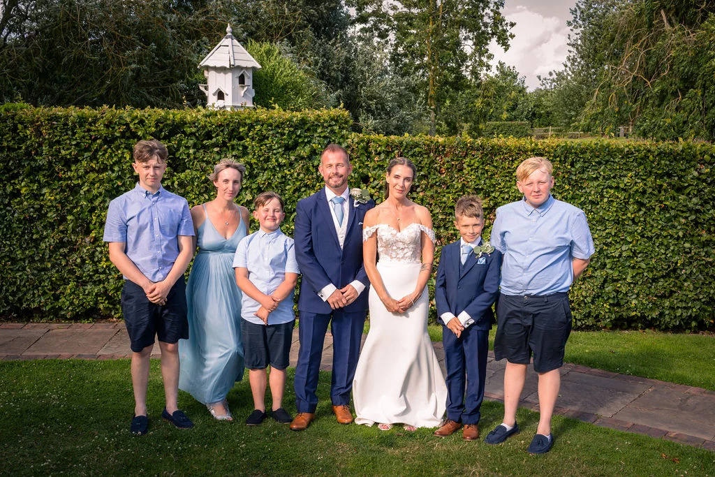 The newlywed couple poses outdoors in a manicured garden alongside their family members, in front of a tall green hedge.