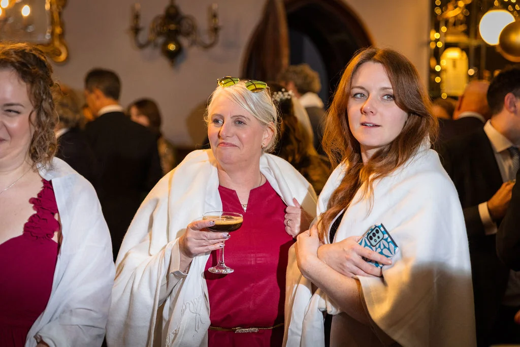 Wedding guests mingle at an evening drinks reception, holding cocktails while draped in white fleece wraps inside a venue.