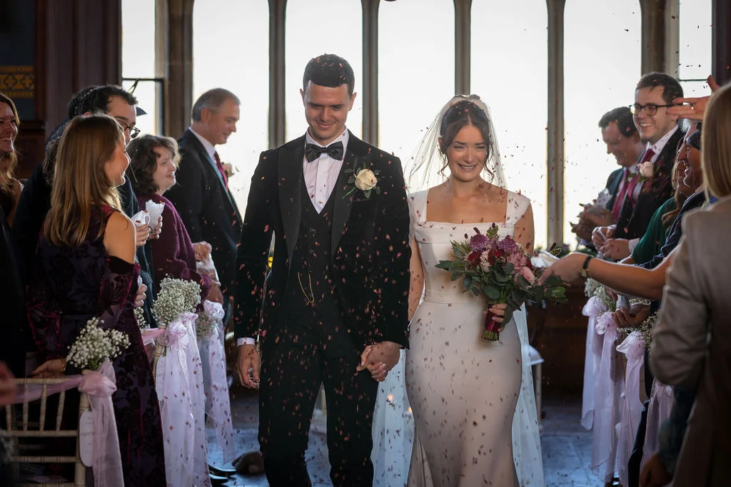 Newlyweds walk hand in hand down the aisle smiling as guests shower them with confetti beneath bright tall windows.