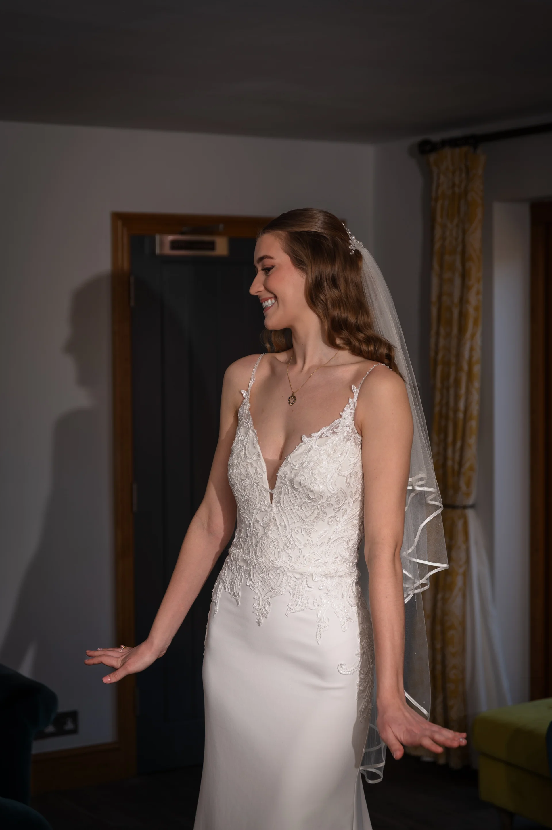 A smiling bride wearing a lace-detailed crepe gown and a veil looks away in profile during her wedding morning preparations.