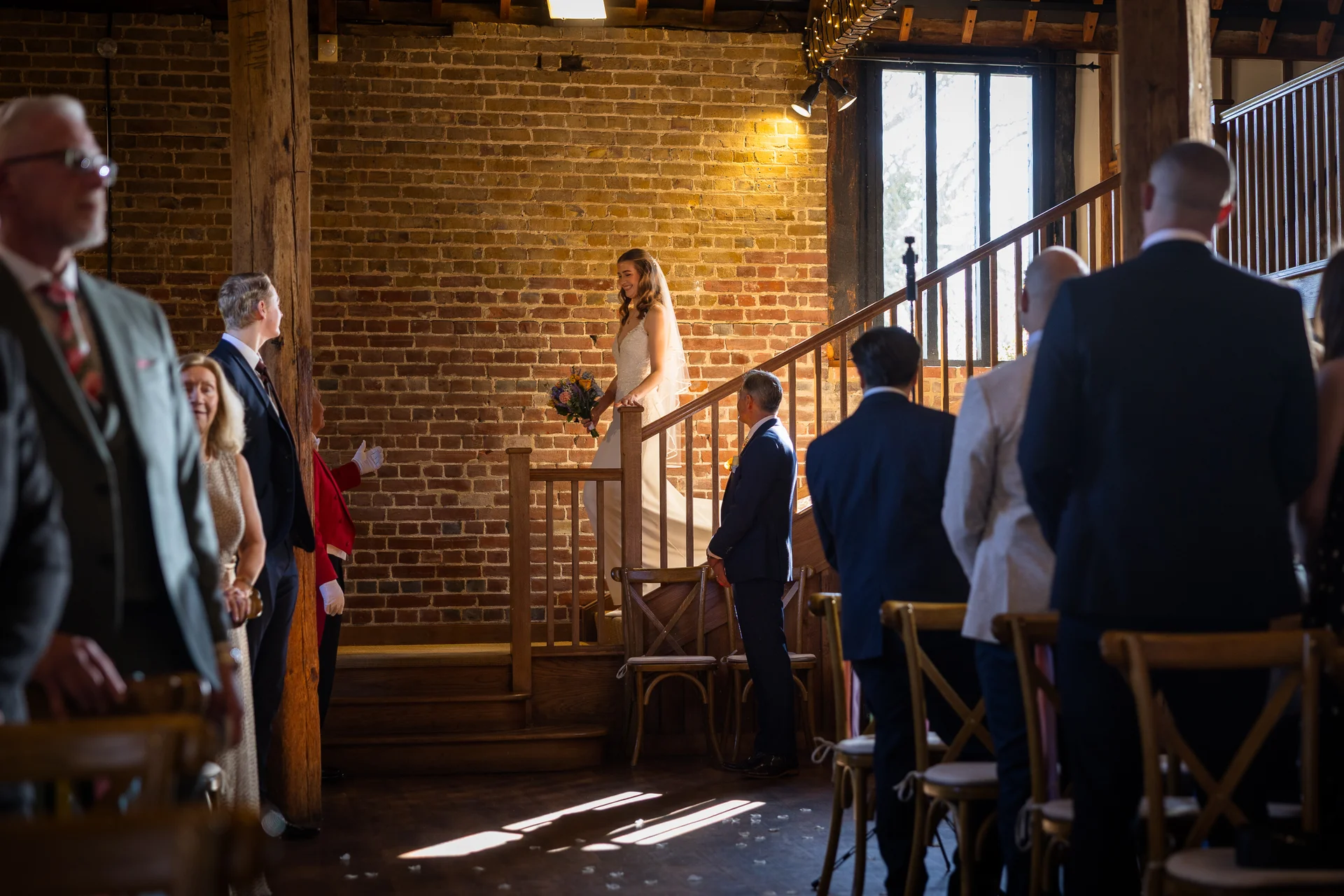 A smiling bride holding a bouquet descends a wooden staircase inside an exposed brick barn while guests look on from the aisle.