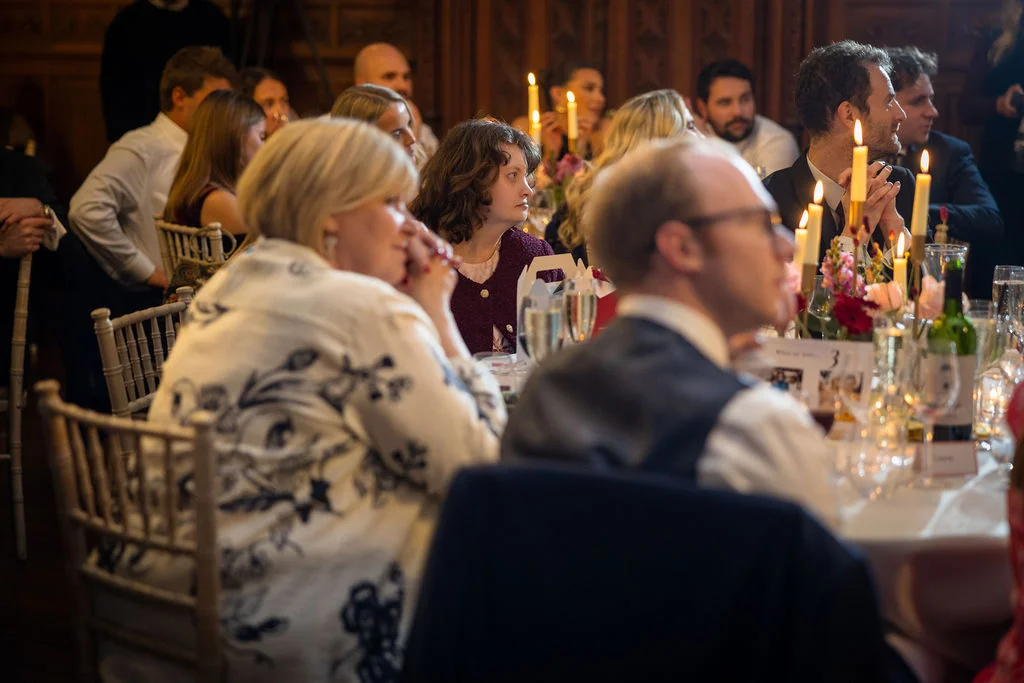 Wedding guests enjoy a candlelit wedding breakfast seated at long tables decorated with floral arrangements and glassware in a wood-panelled venue.