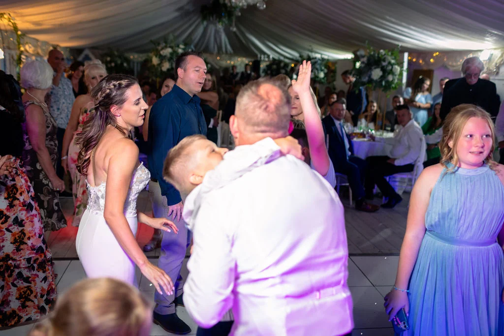 A lively wedding reception in a draped marquee where guests celebrate and dance on the lit dance floor.