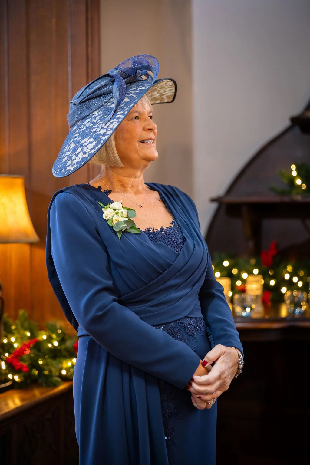 An elegantly dressed mother of the bride wearing a navy blue dress and matching fascinator smiles warmly indoors near twinkling fairy lights.