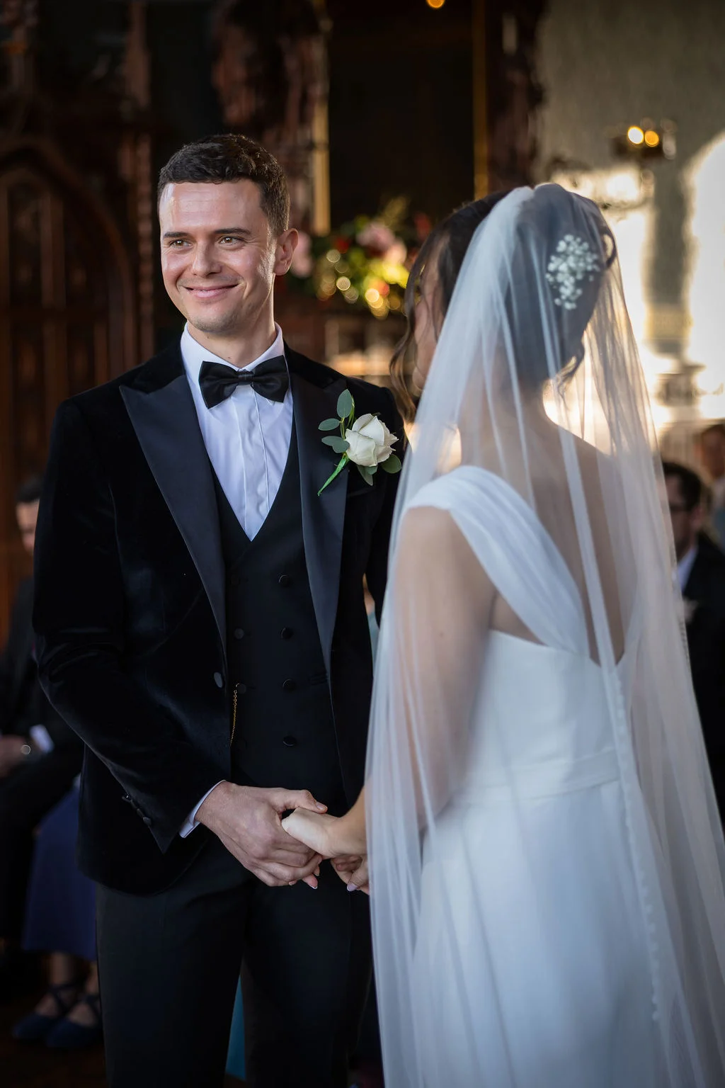 A smiling groom in a black velvet tuxedo and bow tie holds hands with his bride during their wedding ceremony.
