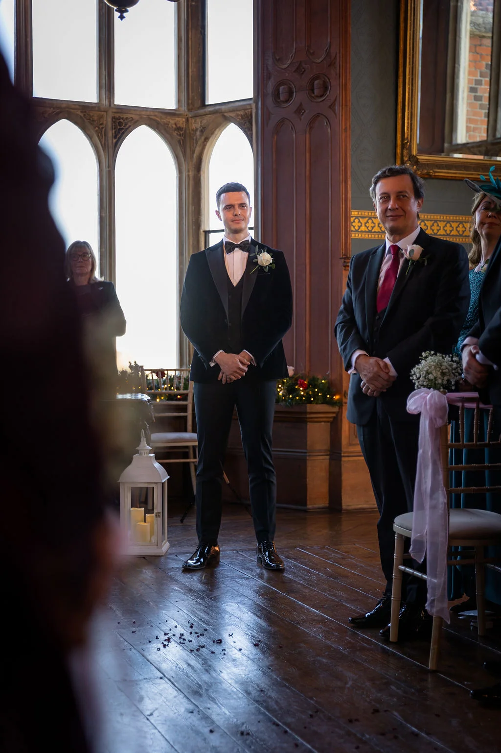 A handsome groom in a black tuxedo and bow tie stands smiling at the altar inside a grand manor house with large arched windows.