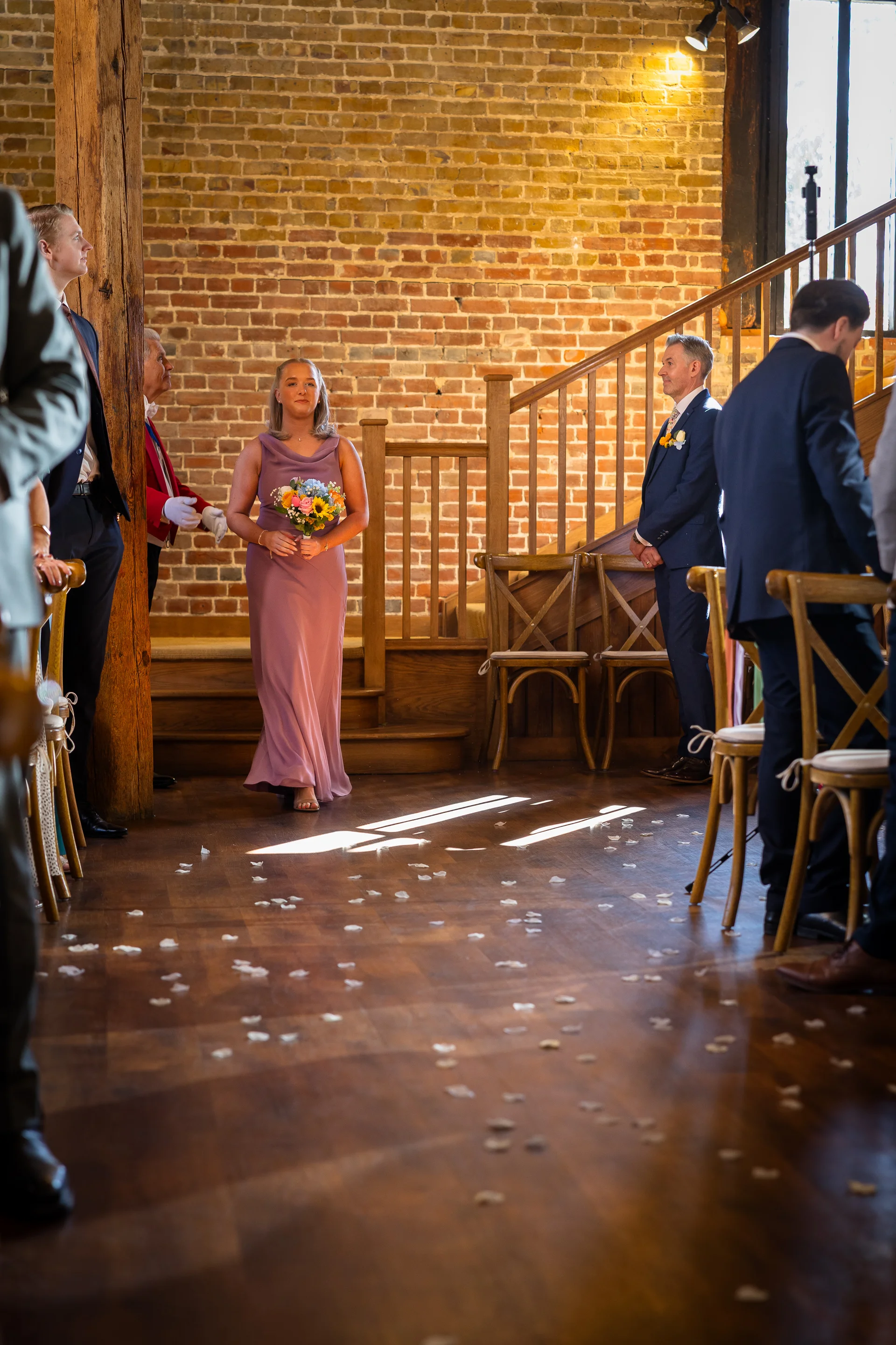 A bridesmaid in a pink dress holding a floral bouquet walks down the aisle inside a rustic brick barn.