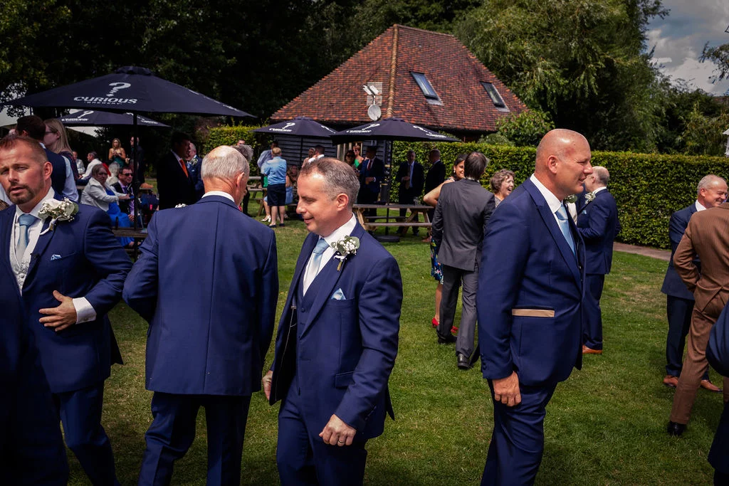 A group of guests and groomsmen in matching navy suits mingle and chat on a lush green lawn during an outdoor wedding reception.