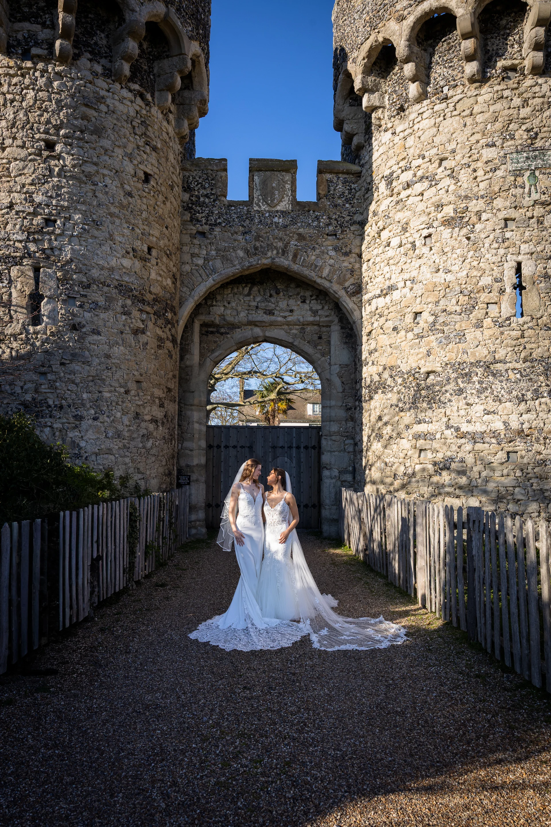 Two brides pose lovingly together in their white lace wedding dresses beneath the grand stone archway of a historic castle gatehouse.