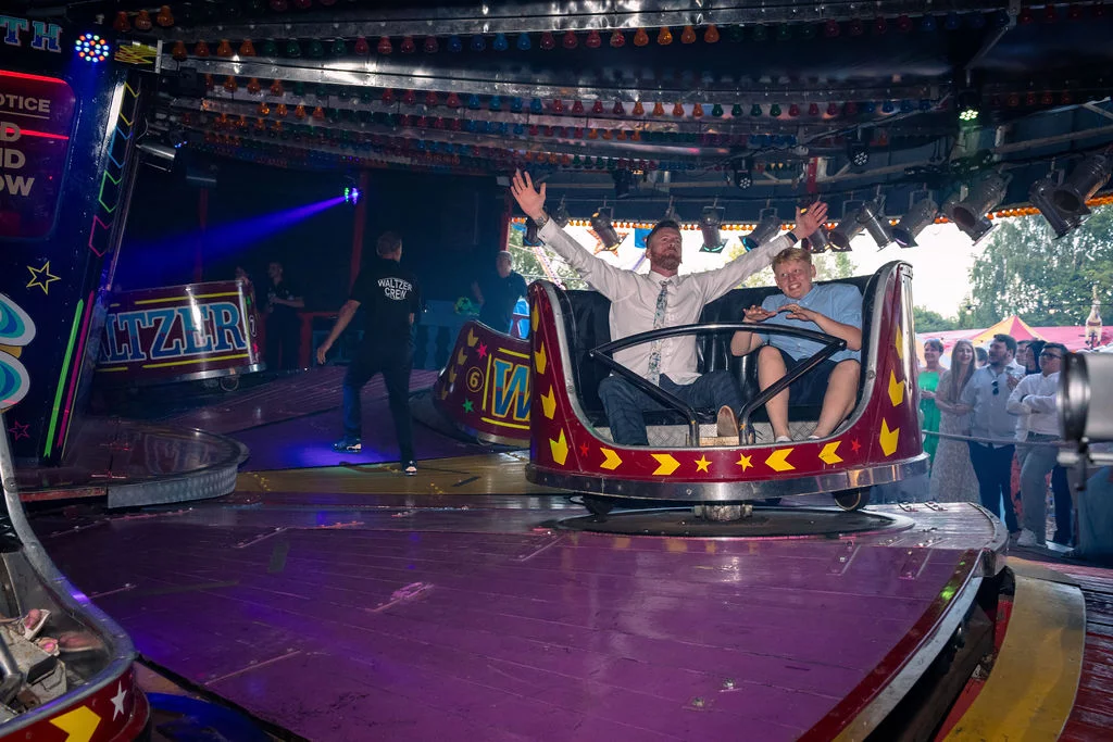 A groom and a guest enjoy a thrilling ride on a neon-lit fairground waltzer at a funfair-themed wedding evening reception.