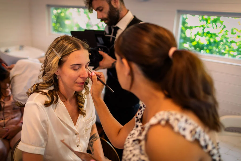 A bride sits calmly in a white robe while a makeup artist applies eye shadow during wedding morning preparations.
