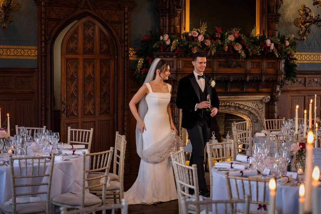 Newlyweds enter a grand wood-panelled dining room decorated with festive floral garlands and glowing candlelight for their wedding reception.