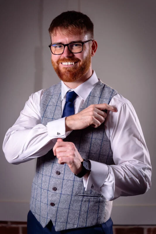 A smiling groom with a red beard and glasses poses in a grey checked tweed waistcoat and a crisp white shirt while adjusting his cufflinks.