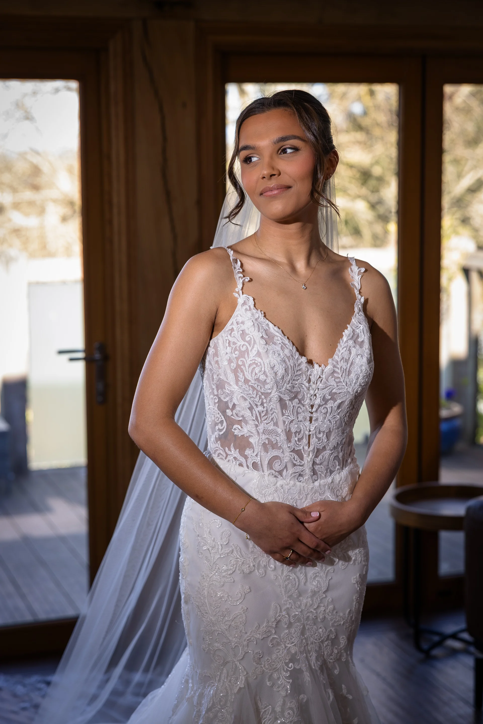 A stunning bride in a lace fit-and-flare wedding dress with spaghetti straps and a sheer corseted bodice stands gracefully indoors near large glass doors.