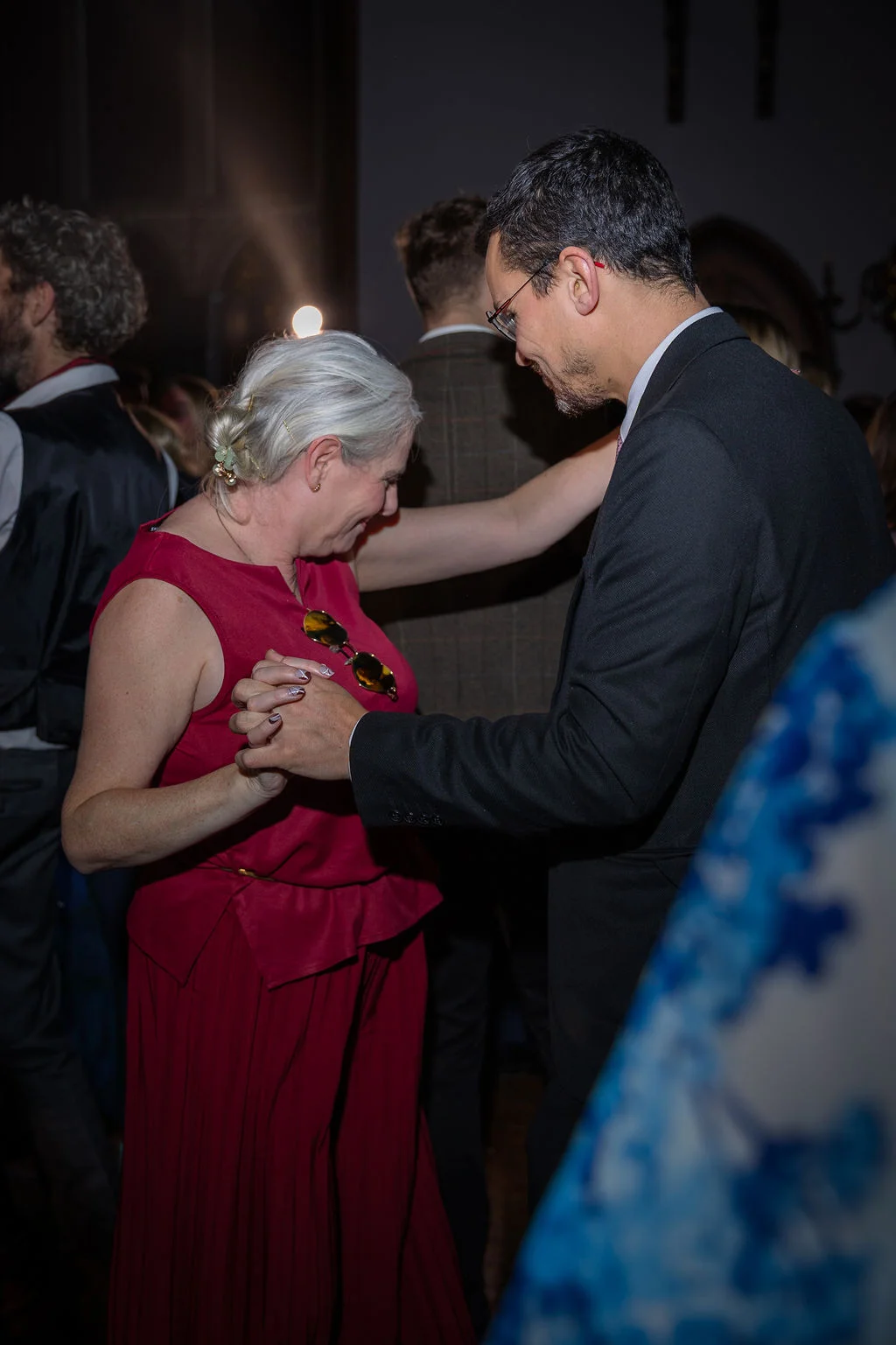 A guest in a red outfit and a man in a black suit share a joyful moment dancing together at an evening wedding reception.