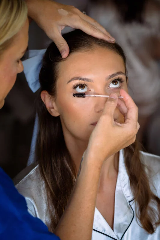 A close-up portrait of a bride having her makeup meticulously applied with a mascara wand during morning bridal preparations.